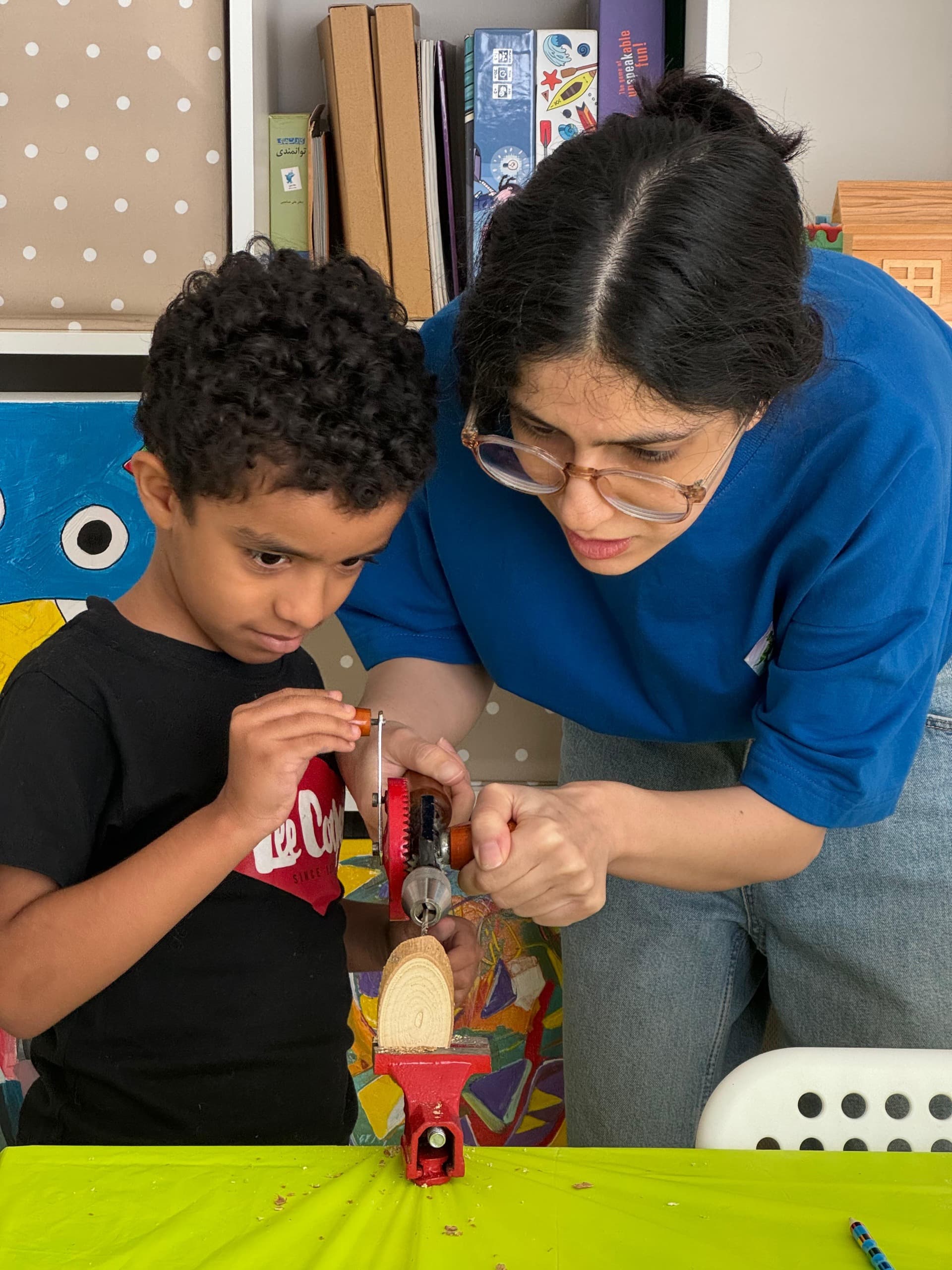 Woman helping a young boy use a hand drill on wood clamped in a vise.